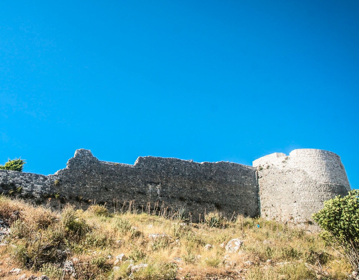 Weathered stone walls and a round tower of Lëkurësi Castle rise above a dry, grassy hillside under a bright blue sky.