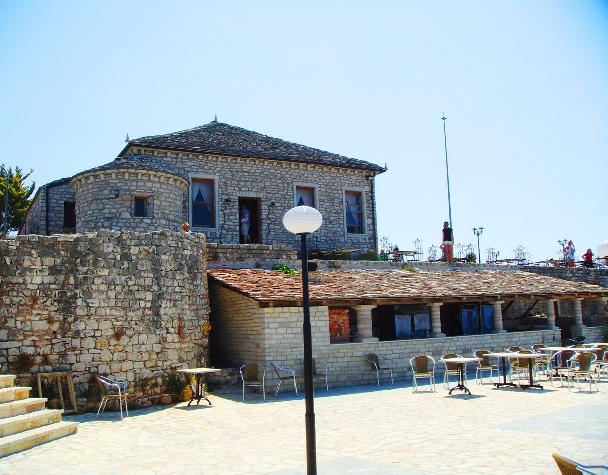 Stone courtyard and terrace at Lëkurësi Castle with rustic buildings, café tables and bright blue sky.