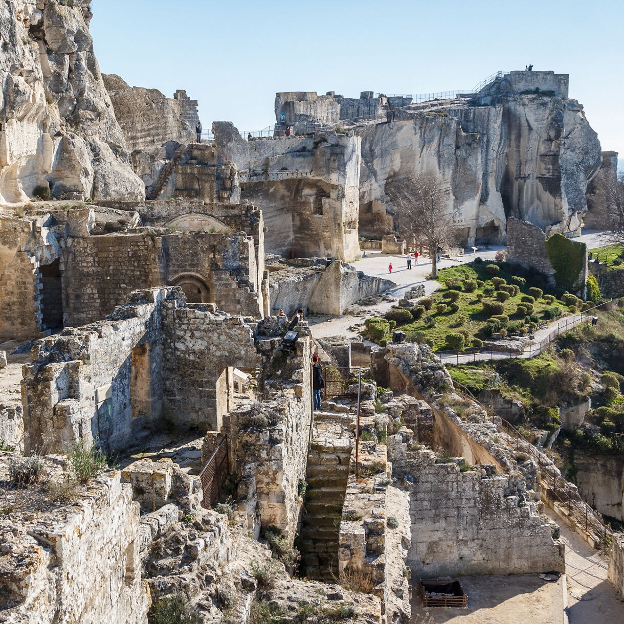 Ancient stone ruins of Les Baux-de-Provence set against rugged cliffs, with pathways and scattered greenery leading visitors through the site.