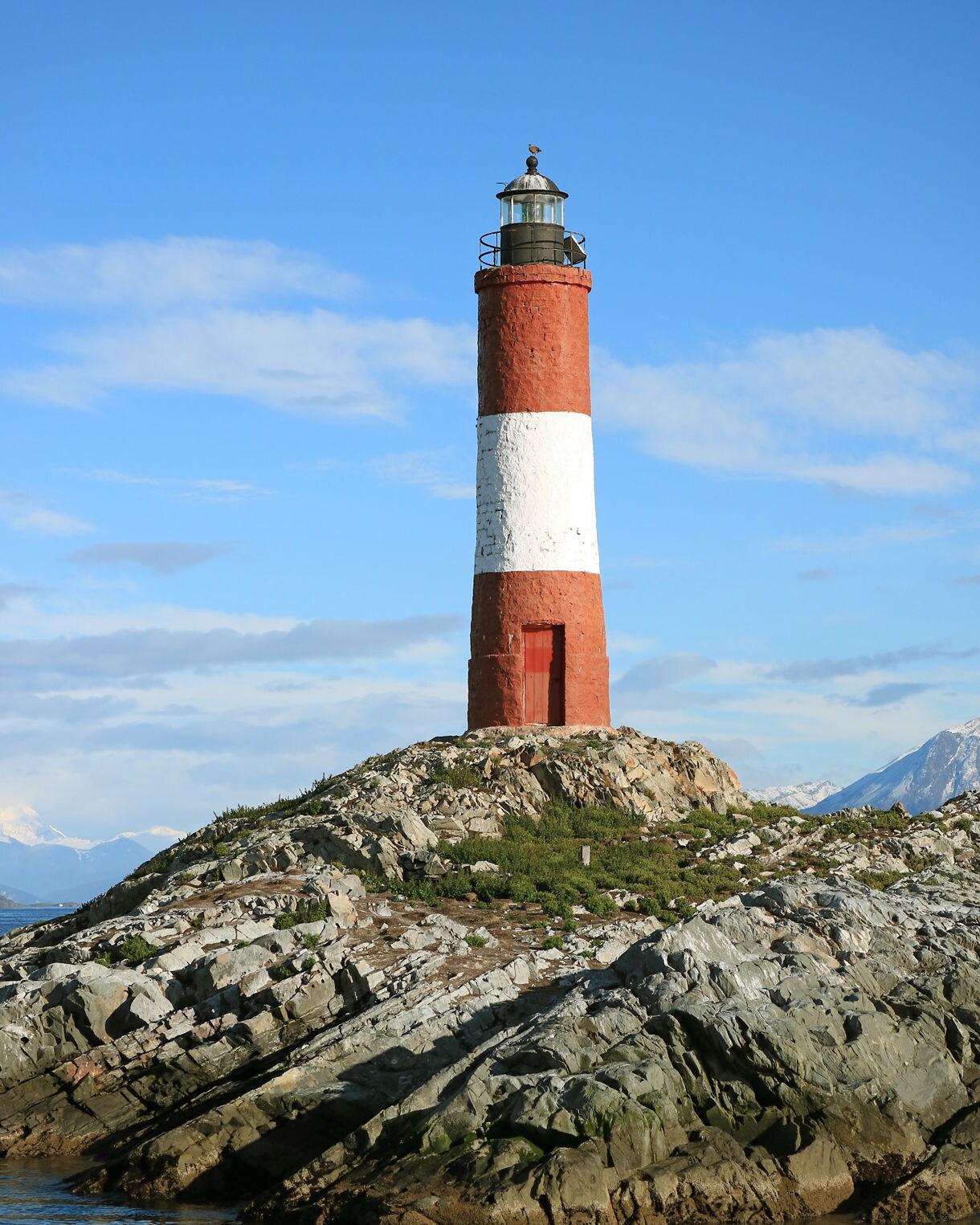 Red and white Les Éclaireurs Lighthouse rising from a rocky islet with calm blue water and distant snowcapped peaks.