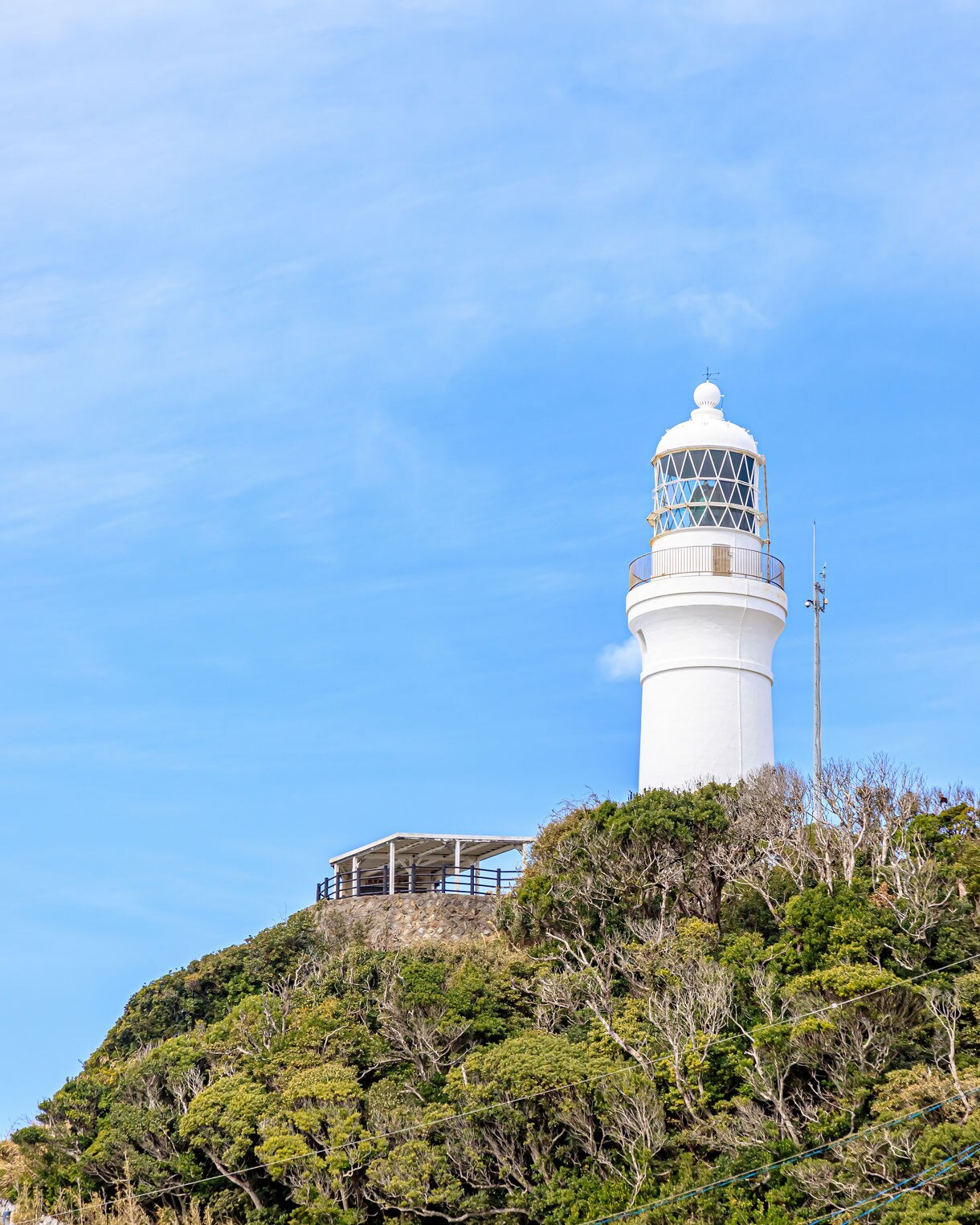 White cylindrical lighthouse with a glass lantern room perched on a green hillside, with a small lookout platform nearby and a clear blue sky overhead.