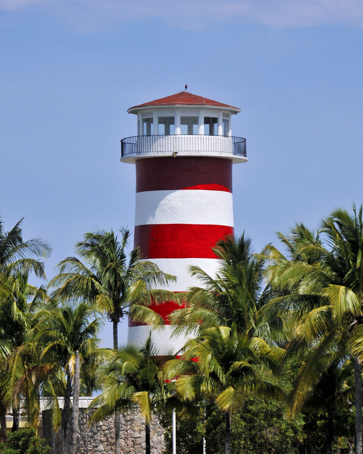 Red and white striped lighthouse surrounded by palm trees under a bright blue sky in Pelican Bay.