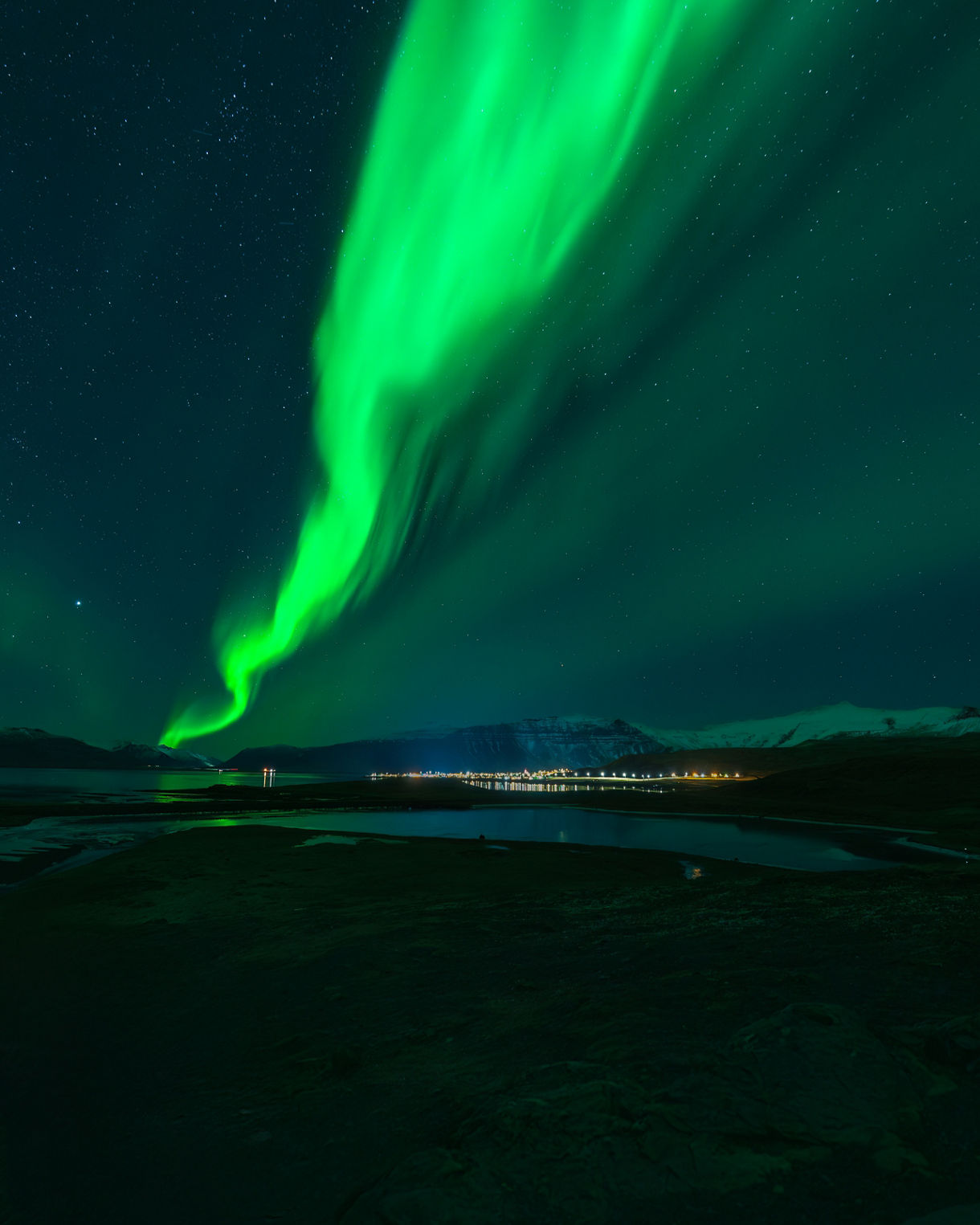 Bright green aurora stretching across a dark night sky above snowy mountains and a distant town, with reflections shimmering on calm water below.
