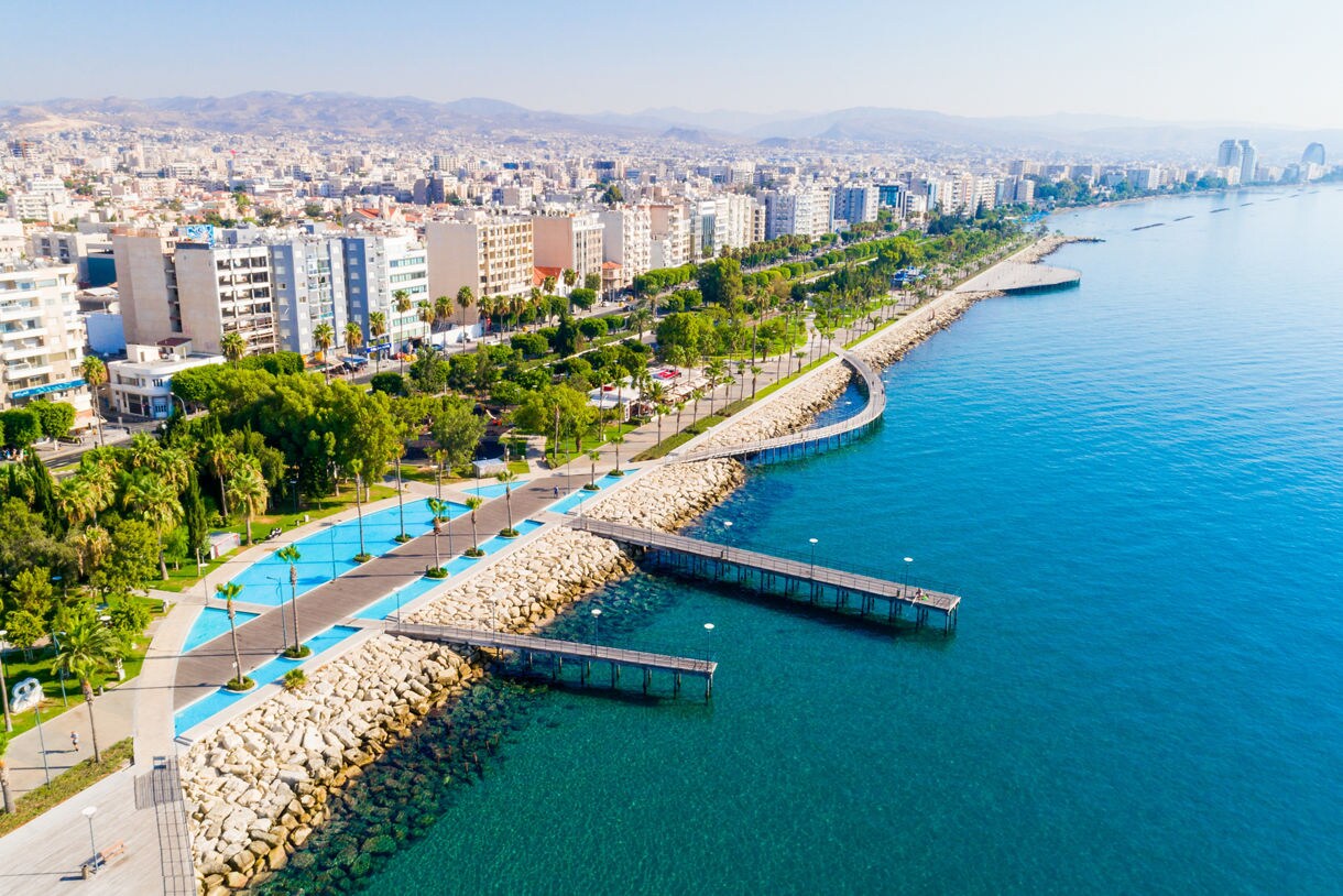 Aerial view of Limassol’s seafront promenade in Cyprus with curved wooden piers, turquoise water, palm-lined paths and a dense cityscape backed by hazy mountains.