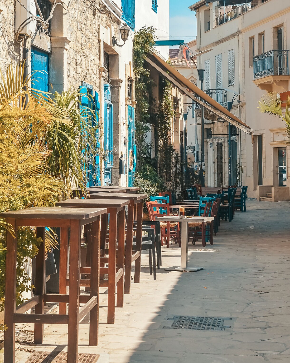Narrow pedestrian street in Limassol with stone buildings, bright blue shutters, outdoor tables and green plants lining the walkway.