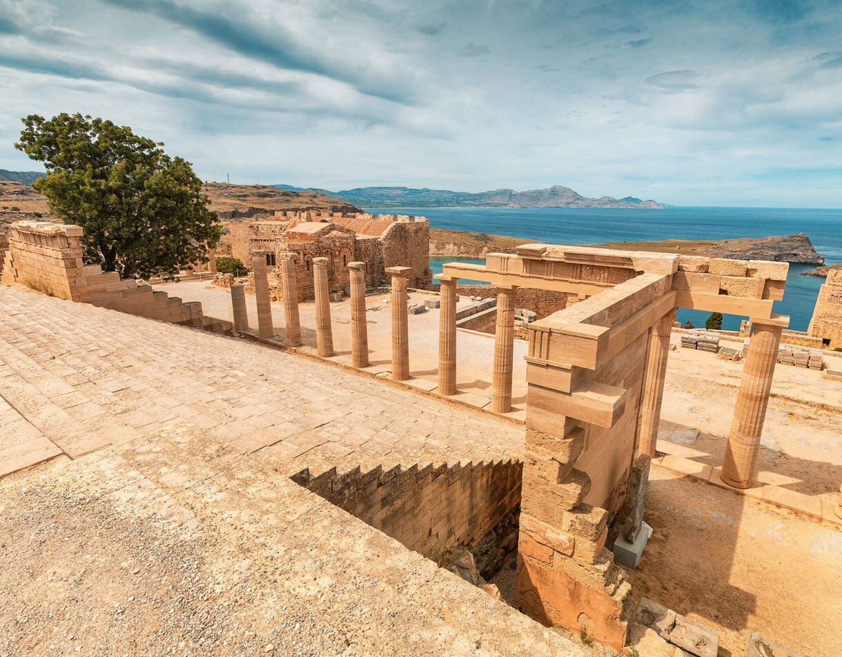 Stone steps and weathered columns of the Lindos Acropolis overlooking turquoise sea and rugged coastline on the island of Rhodes, Greece.