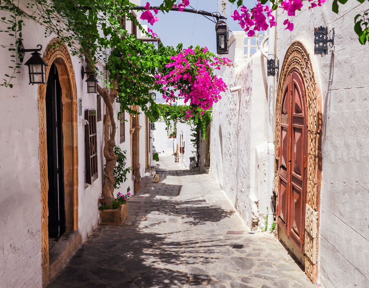 Narrow cobblestone street in Lindos, Greece, lined with whitewashed buildings, wooden doors and bright pink bougainvillea flowers overhead.