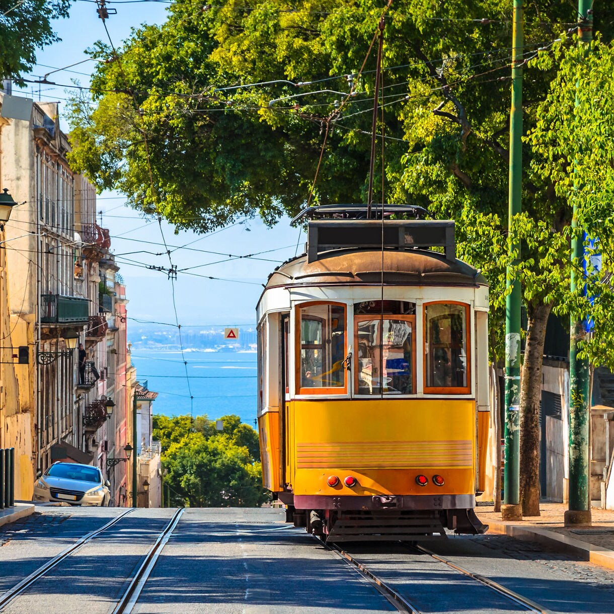 A classic yellow tram travels down a steep Lisbon street lined with old buildings and green trees, with the blue Atlantic in the distance.