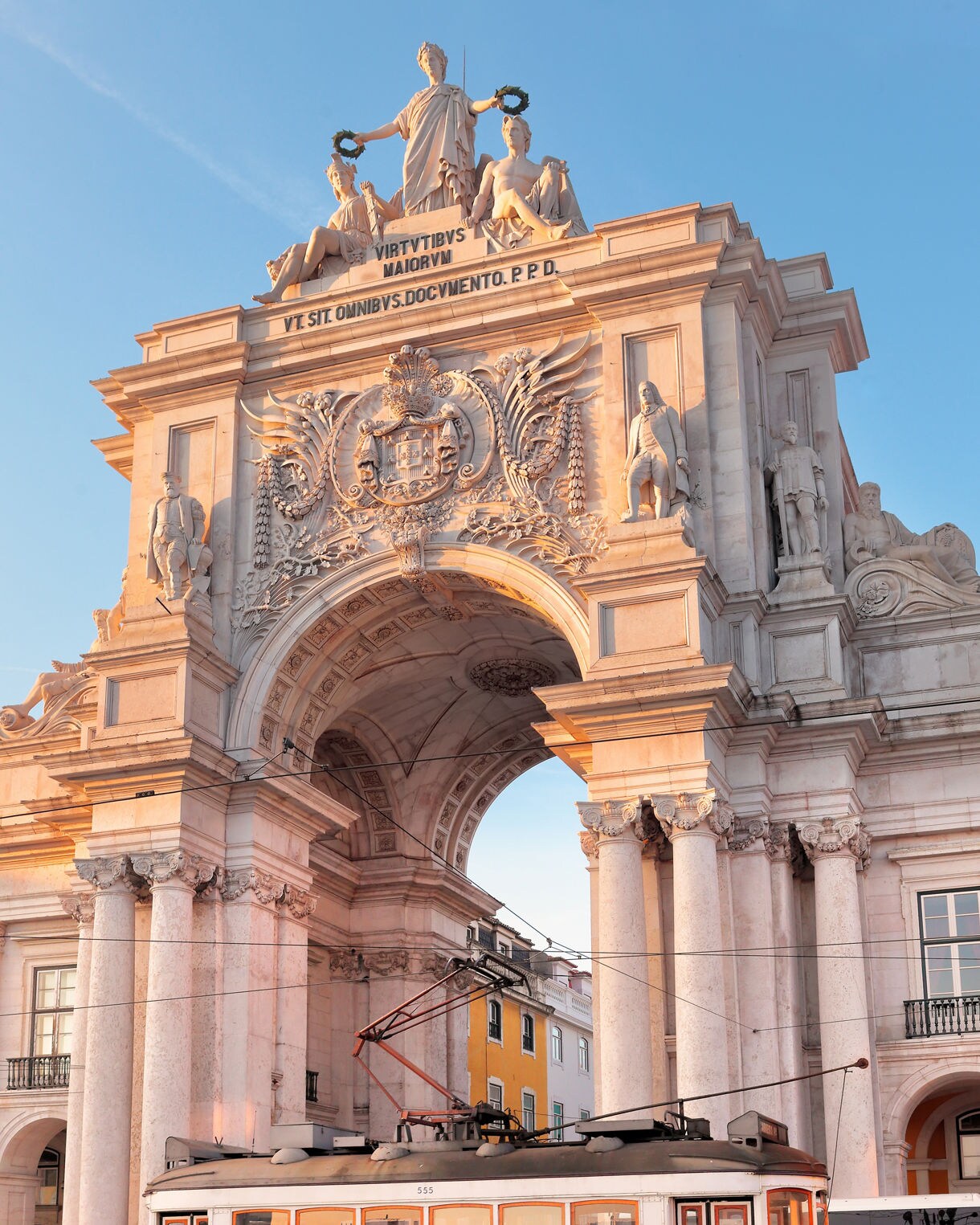 Yellow tram moving under the ornate Arco da Rua Augusta in Lisbon, framed by marble columns and statues.