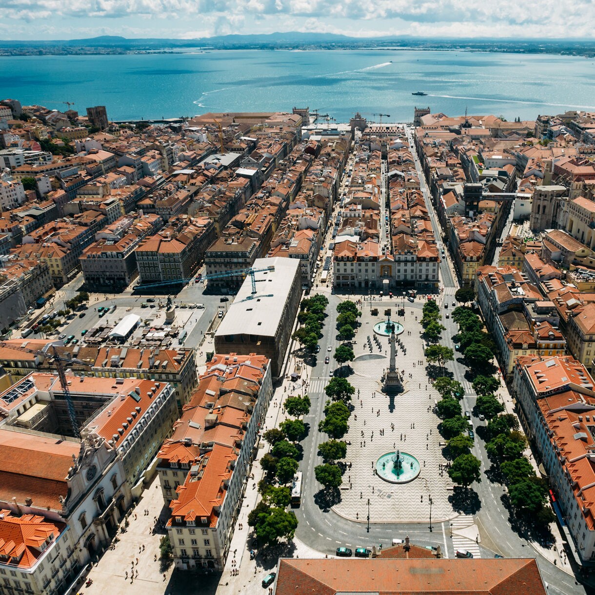 Aerial view of Rossio Square in Lisbon, Portugal, with patterned stone pavement, fountains and surrounding buildings leading toward the Tagus River.