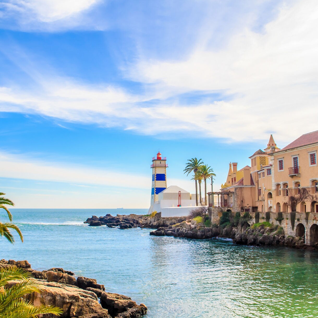 Scenic view of Cascais, Portugal, featuring a blue-and-white striped lighthouse, palm trees and seaside villas along rocky shores under a bright sky.