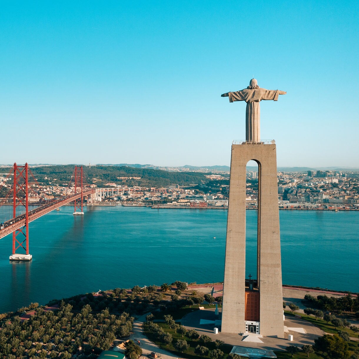 Aerial view of Lisbon’s Cristo Rei monument with arms outstretched, facing the red 25 de Abril suspension bridge spanning the Tagus River.