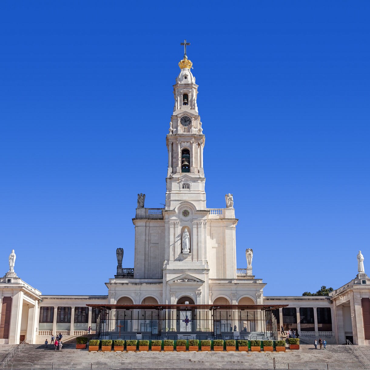 The grand white façade and bell tower of the Sanctuary of Fátima in Portugal, set against a clear blue sky with visitors walking near the entrance.