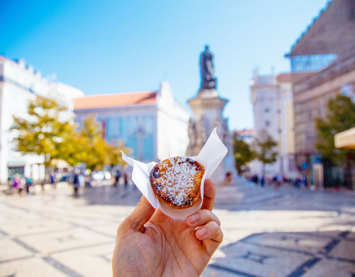 Hand holding a pastel de nata pastry with powdered sugar in a Lisbon square, statue and historic buildings blurred in the background.
