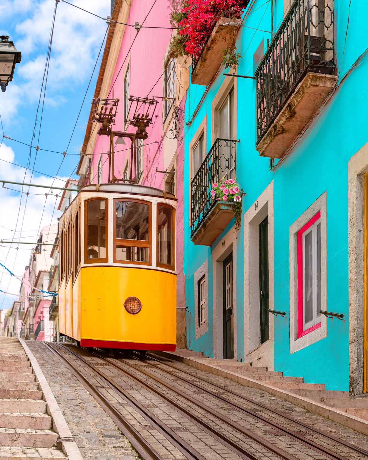Bright yellow Lisbon tram on a steep cobbled street lined with turquoise, pink and green buildings decorated with flowers.