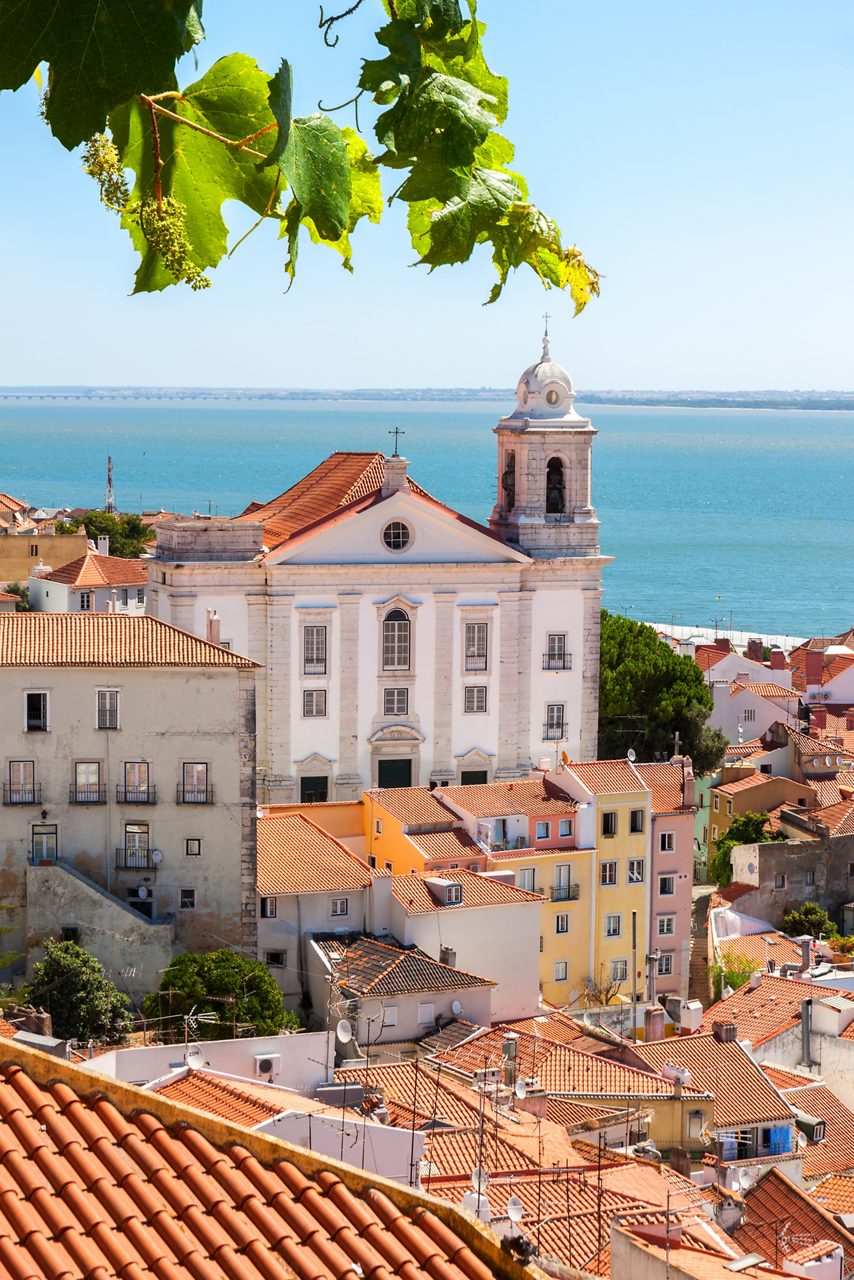 White Igreja de São Vicente de Fora with red-tiled houses in Alfama, overlooking the blue sea.