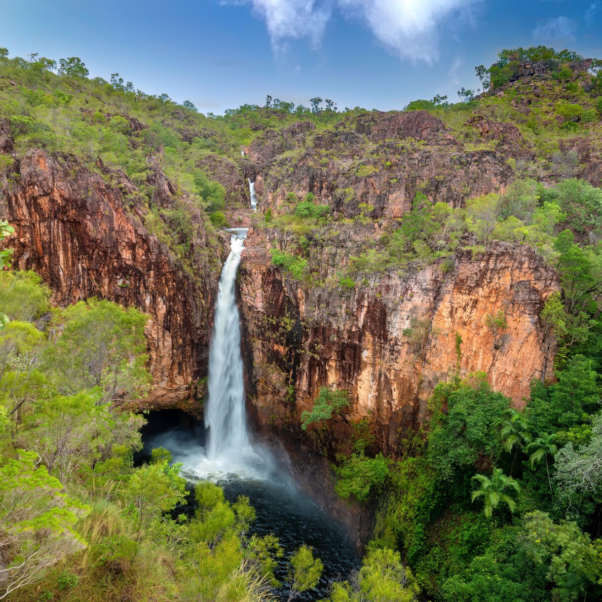 Tall waterfall cascading into a deep rock pool amid red cliffs and dense green vegetation in Litchfield National Park, Australia.
