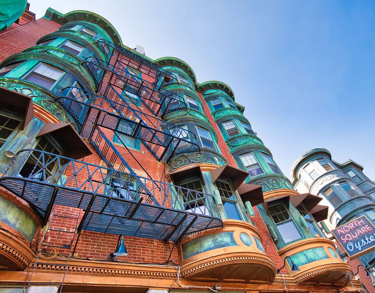Upward view of a historic Boston North End building with green-patina copper bay windows, red brick walls and metal fire escapes against a bright blue sky.