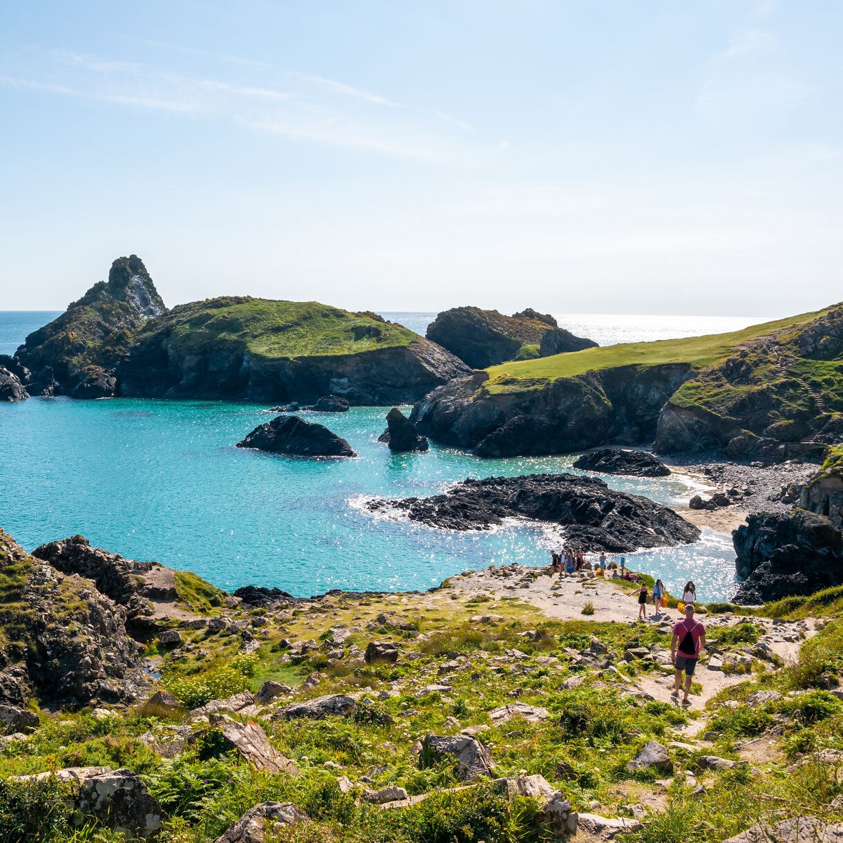 People walking along a rocky path leading to a turquoise cove surrounded by green cliffs and jagged rocks on Cornwall’s Lizard Peninsula.
