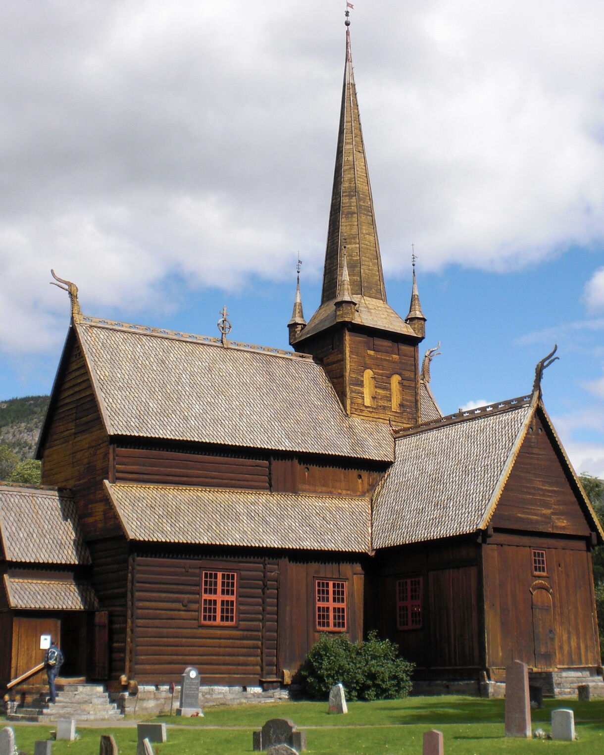 Lom Stave Church, a large wooden medieval church with steep roofs, carved dragon-head details and a tall central spire, surrounded by a graveyard and trees under a cloudy sky.