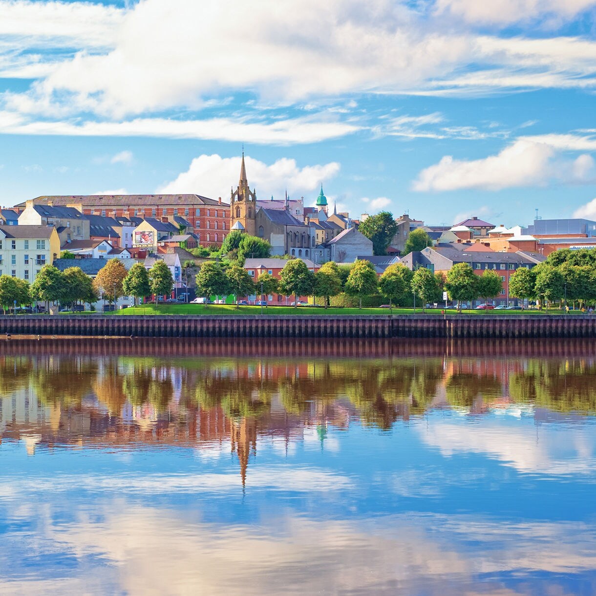 Panoramic view of Londonderry with historic churches, colorful buildings and their reflections in the river under a partly cloudy sky.