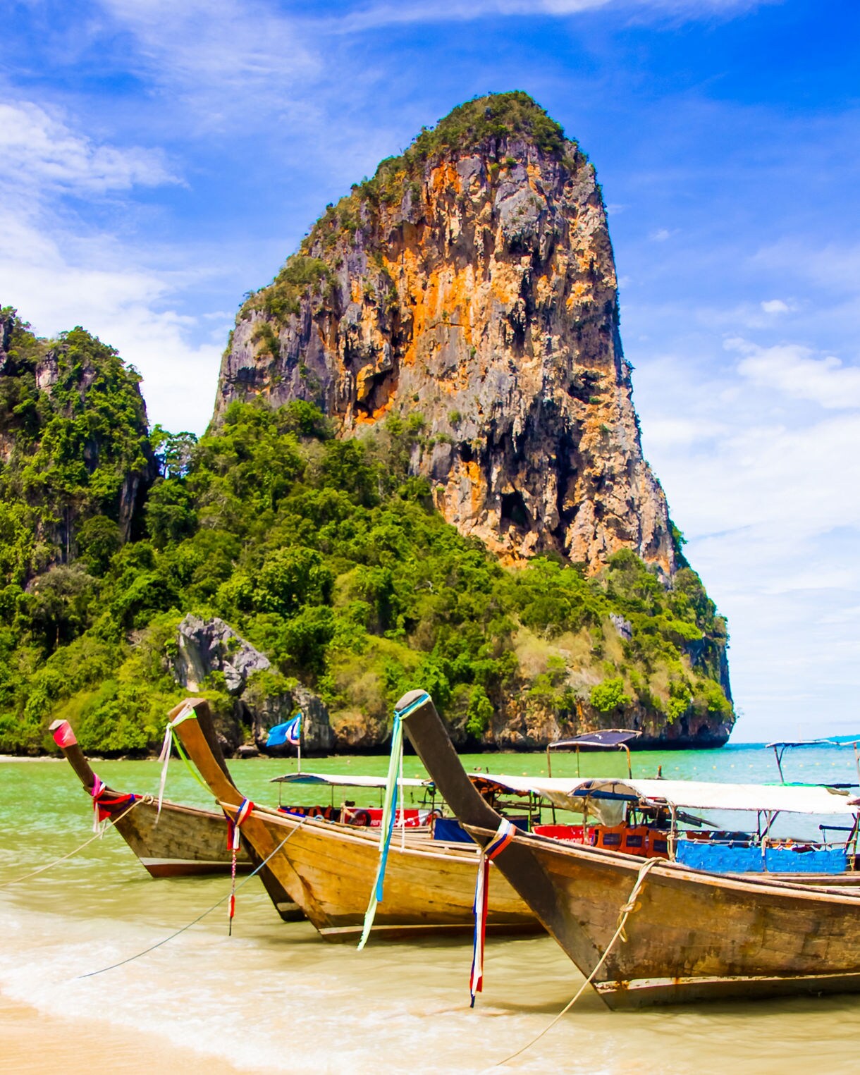 Traditional wooden longtail boats rest in shallow turquoise water with a tall, rugged limestone cliff rising behind them.