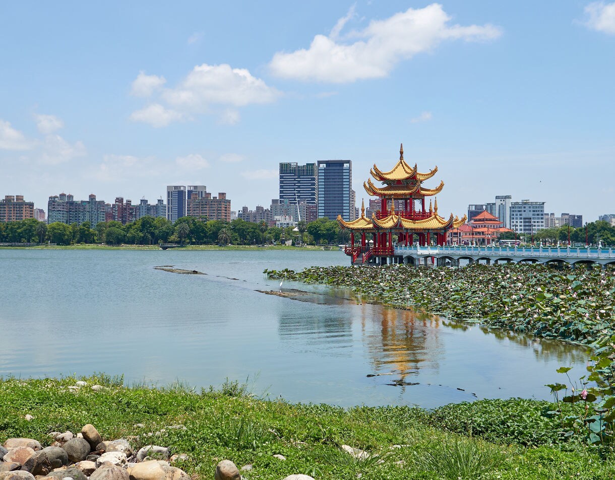 Scenic view of Lotus Lake in Kaohsiung, Taiwan, featuring a traditional red-and-gold Chinese pavilion surrounded by lotus plants with city buildings in the background.