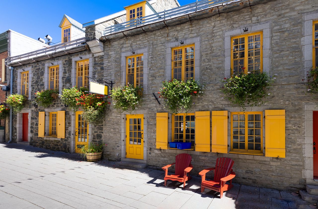 Stone building in Québec City’s Lower Town with yellow doors and shutters, hanging flower baskets and two red chairs set on a sunny cobblestone street.