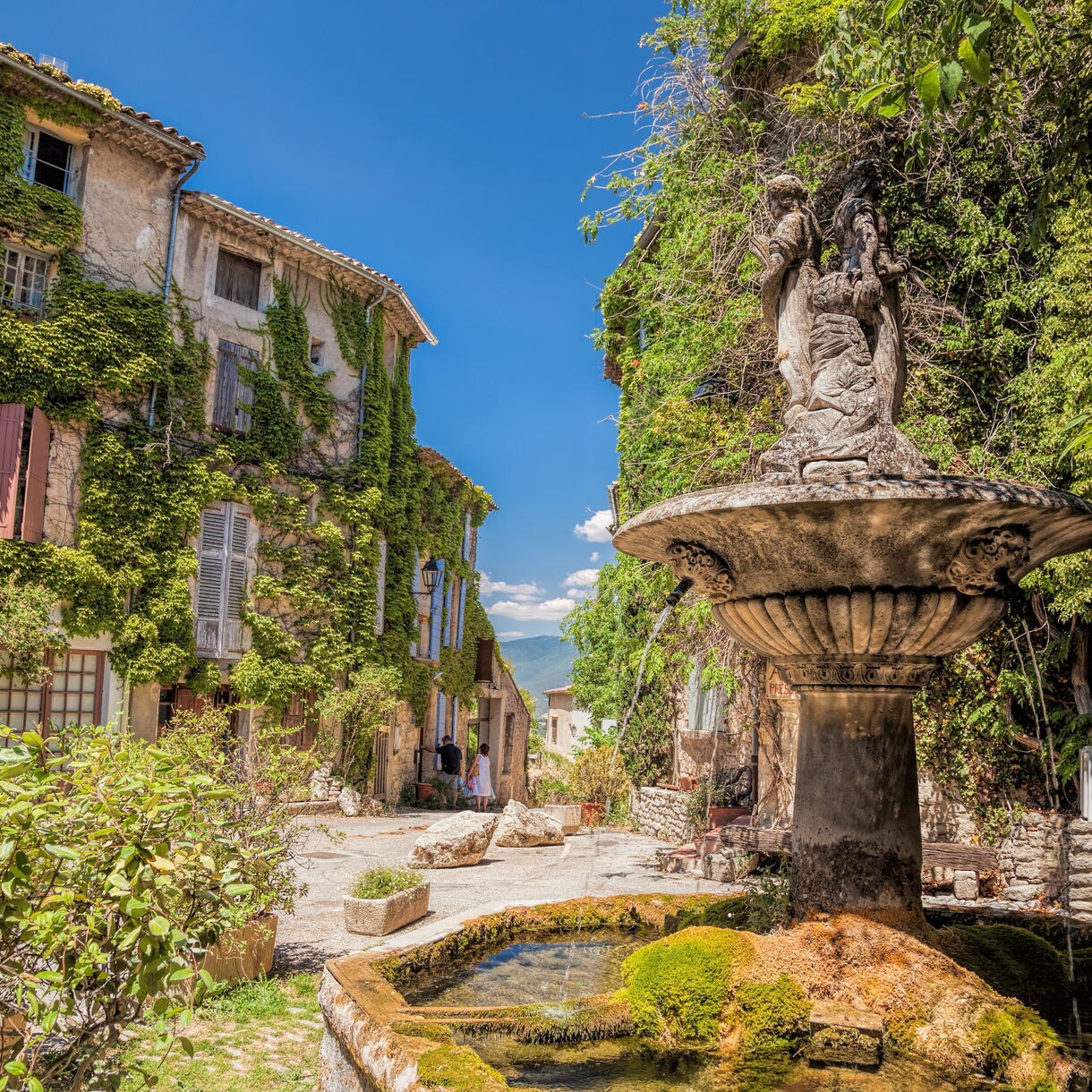 A sunlit square in a Luberon village with ivy-covered stone houses, colorful shutters and an old mossy fountain in the foreground.
