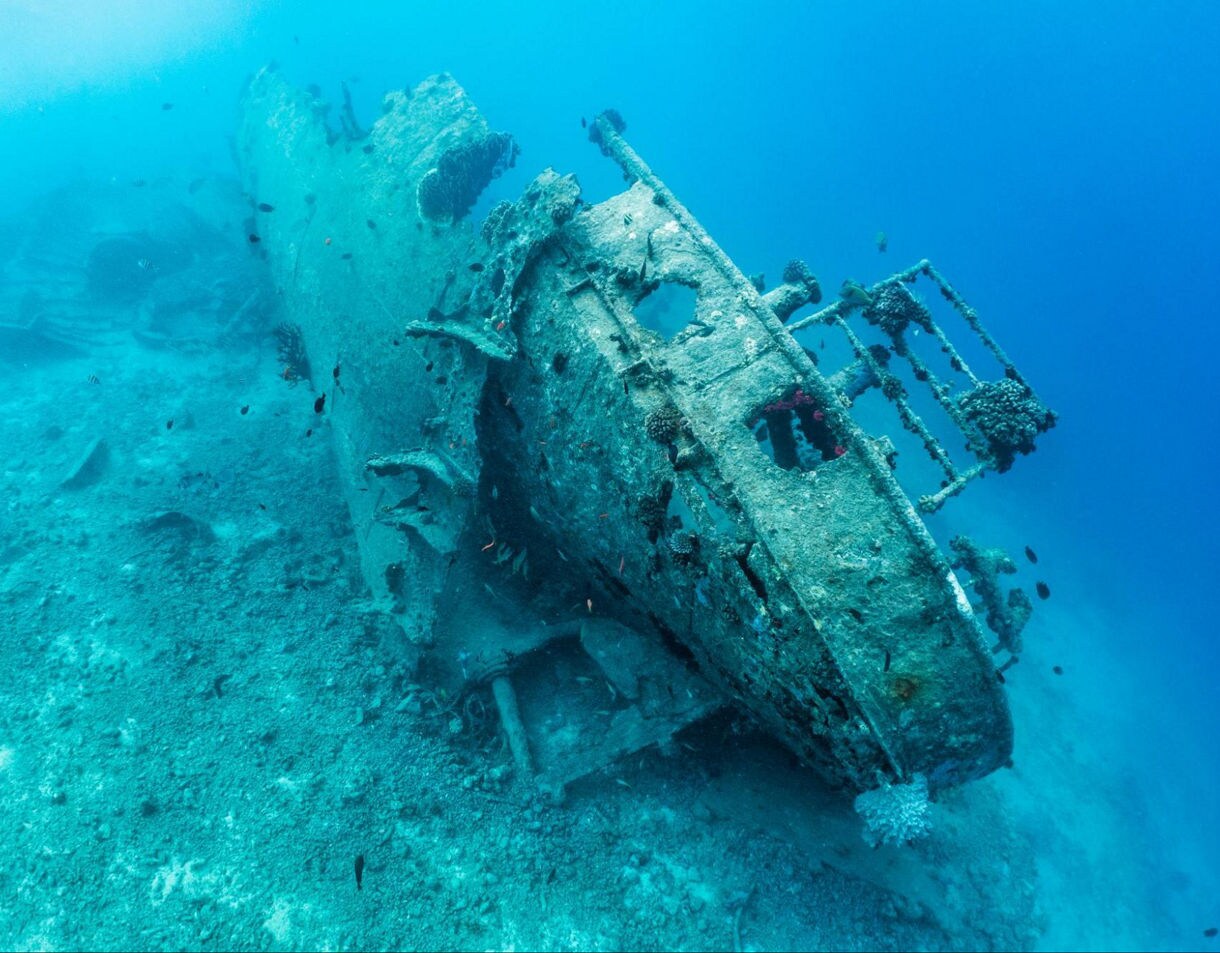Underwater view of a large rusted shipwreck resting on the ocean floor, covered with coral and surrounded by small tropical fish in clear blue water.