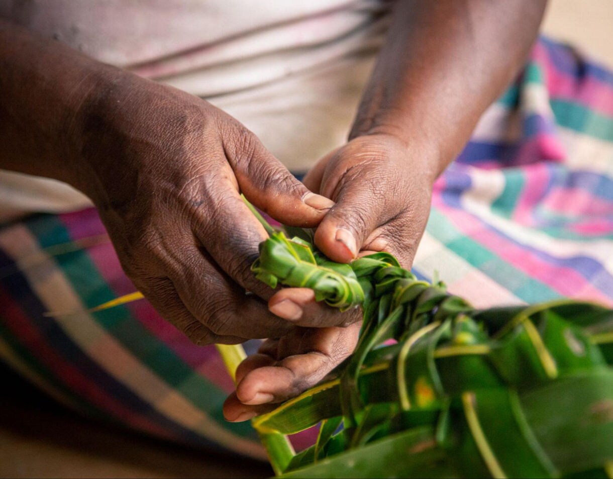 Close-up of a person’s hands weaving green palm leaves into a braid, with a colorful woven cloth visible in the background.