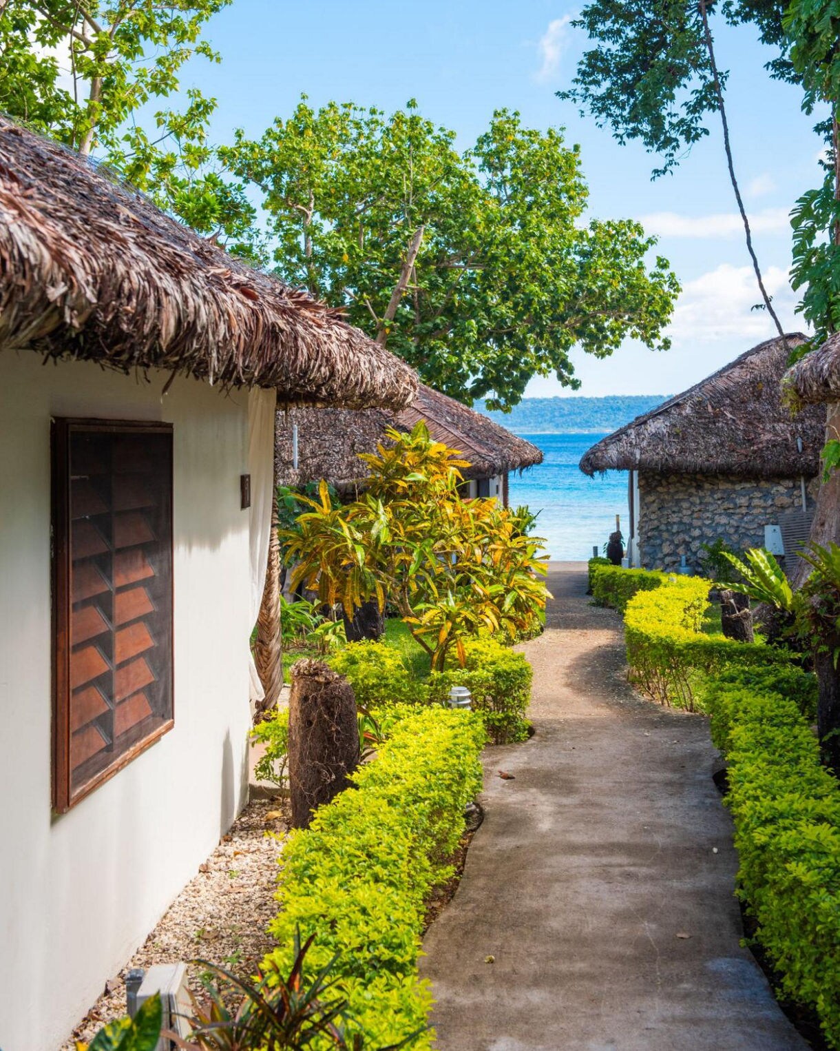 A tropical walkway lined with bright green hedges leading between thatched-roof bungalows toward a blue ocean, with lush trees overhead.