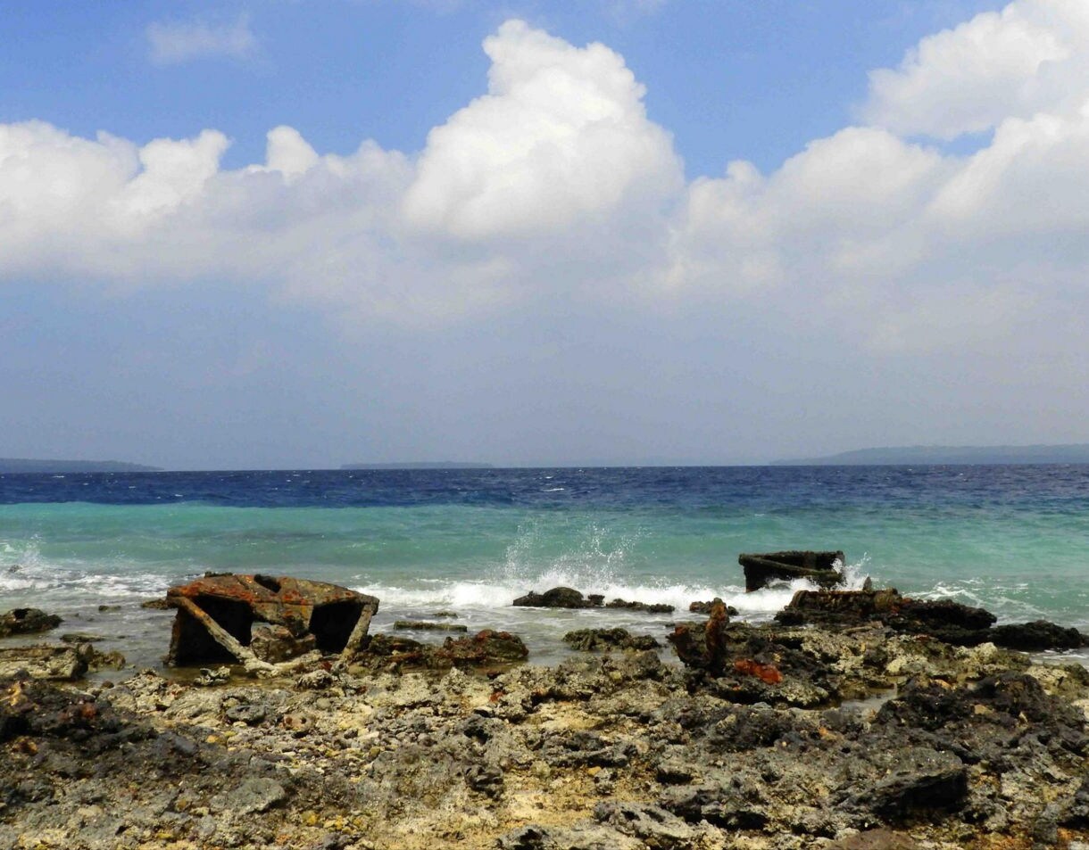 Rusted metal remnants from WWII scattered along a rocky shoreline with turquoise waves and distant land on the horizon.