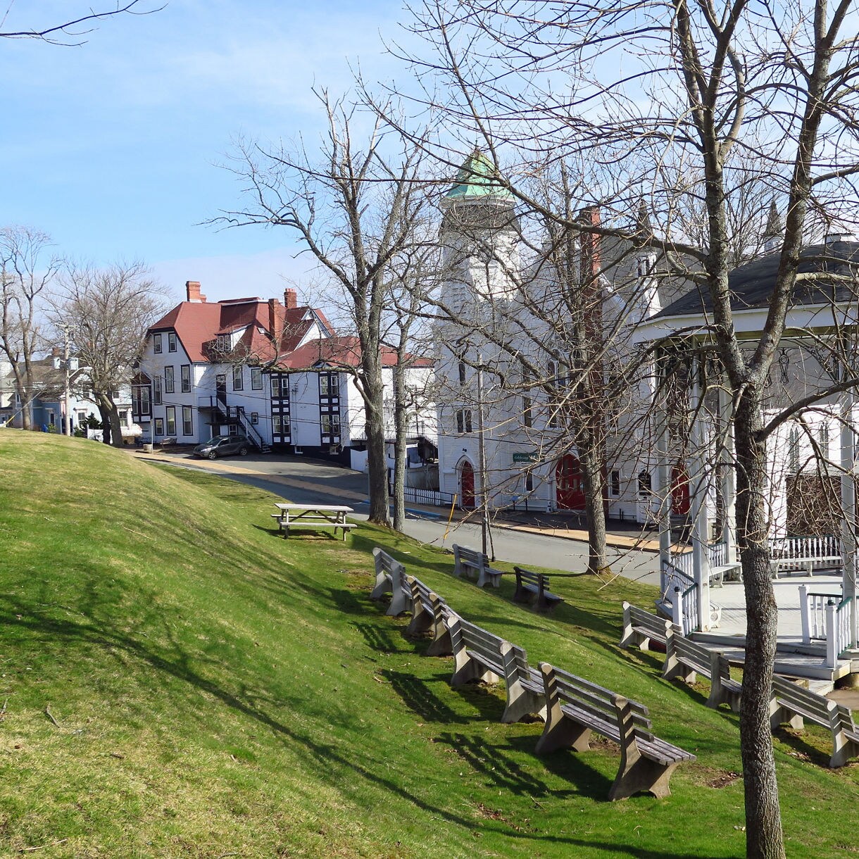 Sloping grassy park in Lunenburg with wooden benches, a white bandstand and historic red-roofed buildings lining the street.