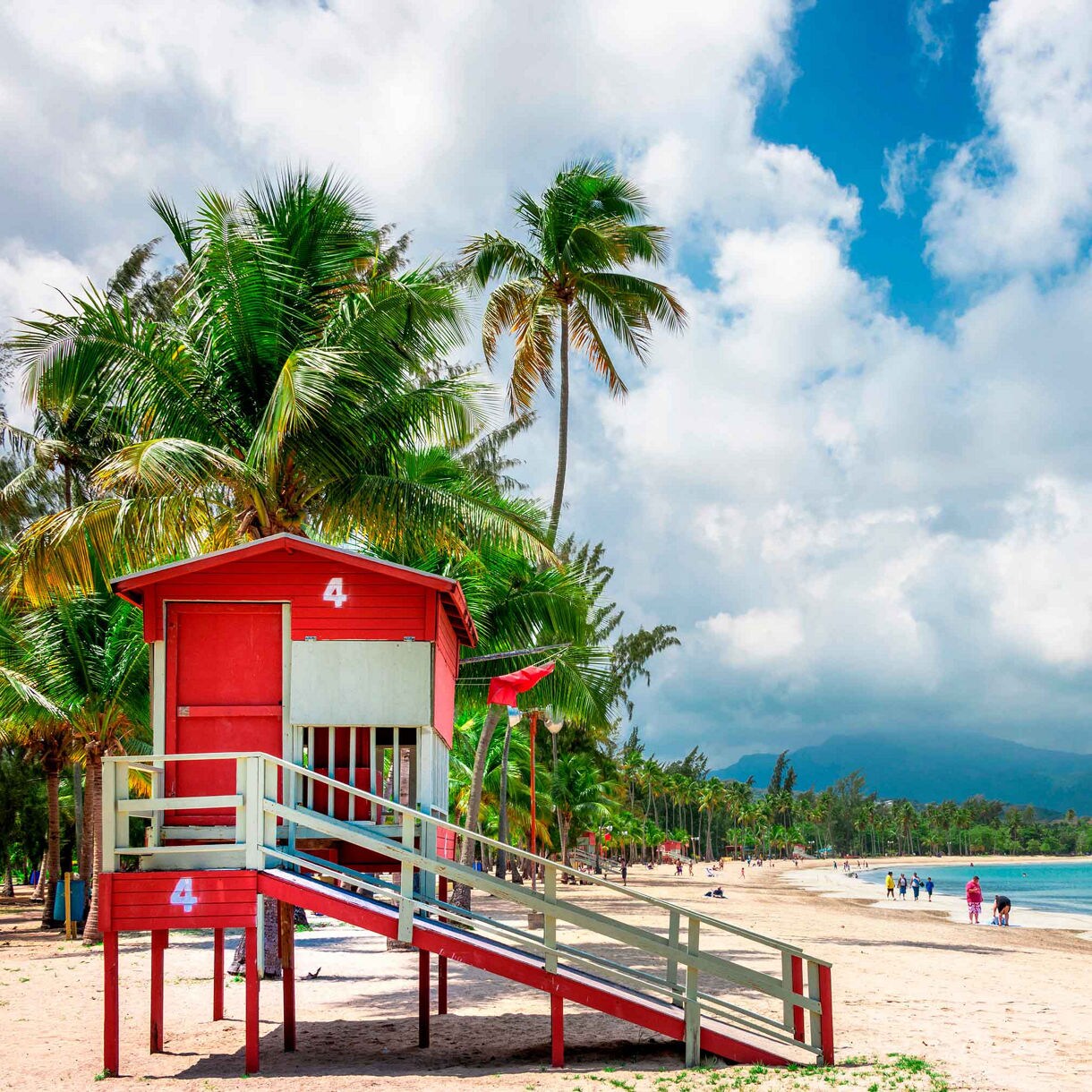 Red lifeguard tower on Luquillo Beach in Puerto Rico, with palm trees, beachgoers and distant mountains under a partly cloudy sky.