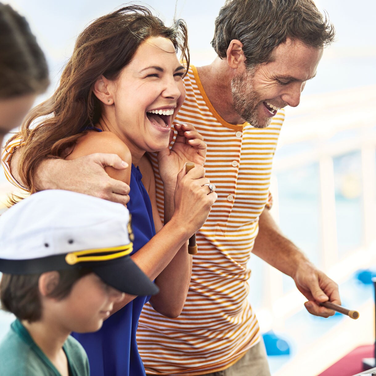 A group of people, including adults and children, are gathered on the deck of a cruise ship, engaging in an interactive activity. The setting is bright and sunny, with a relaxed and cheerful atmosphere. The adults appear to be guiding the children, who are focused on the activity. Nautical elements and casual summer clothing are visible, enhancing the vacation vibe. No visible text or numbers are present in the image.