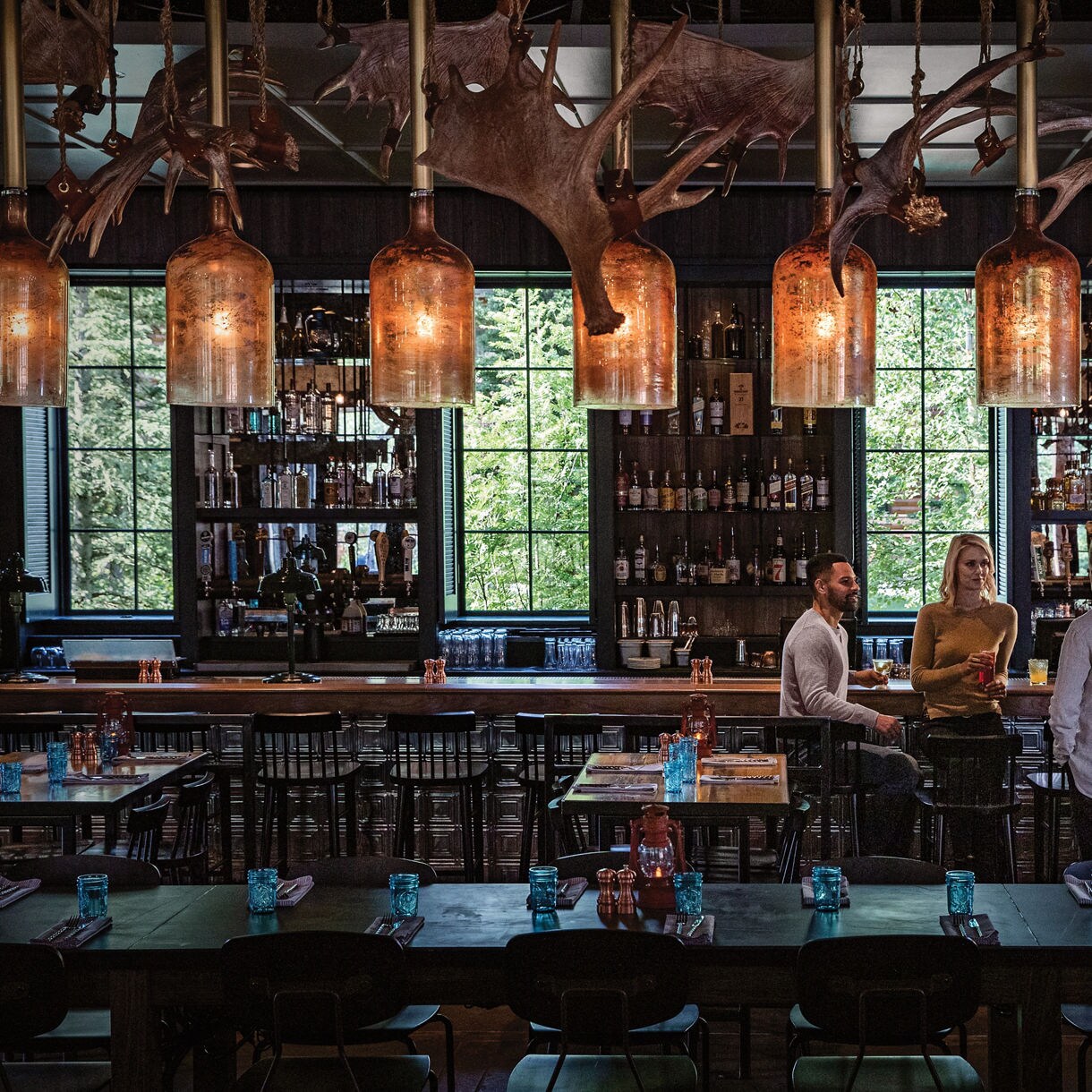 Four adults are gathered around a table in a stylish bar, engaged in conversation. The setting features large windows, shelves of bottles, and prominent antler decorations hanging from the ceiling. Warm lighting and blue glassware create a cozy, inviting mood. The overall palette is earthy and atmospheric, with a mix of wood and metal elements.