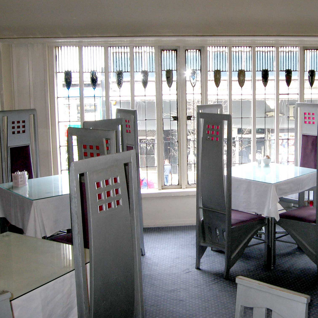 Interior of the Willow Tea Rooms in Glasgow featuring tall, geometric high-backed chairs and stained glass windows in Mackintosh’s signature Art Nouveau style.