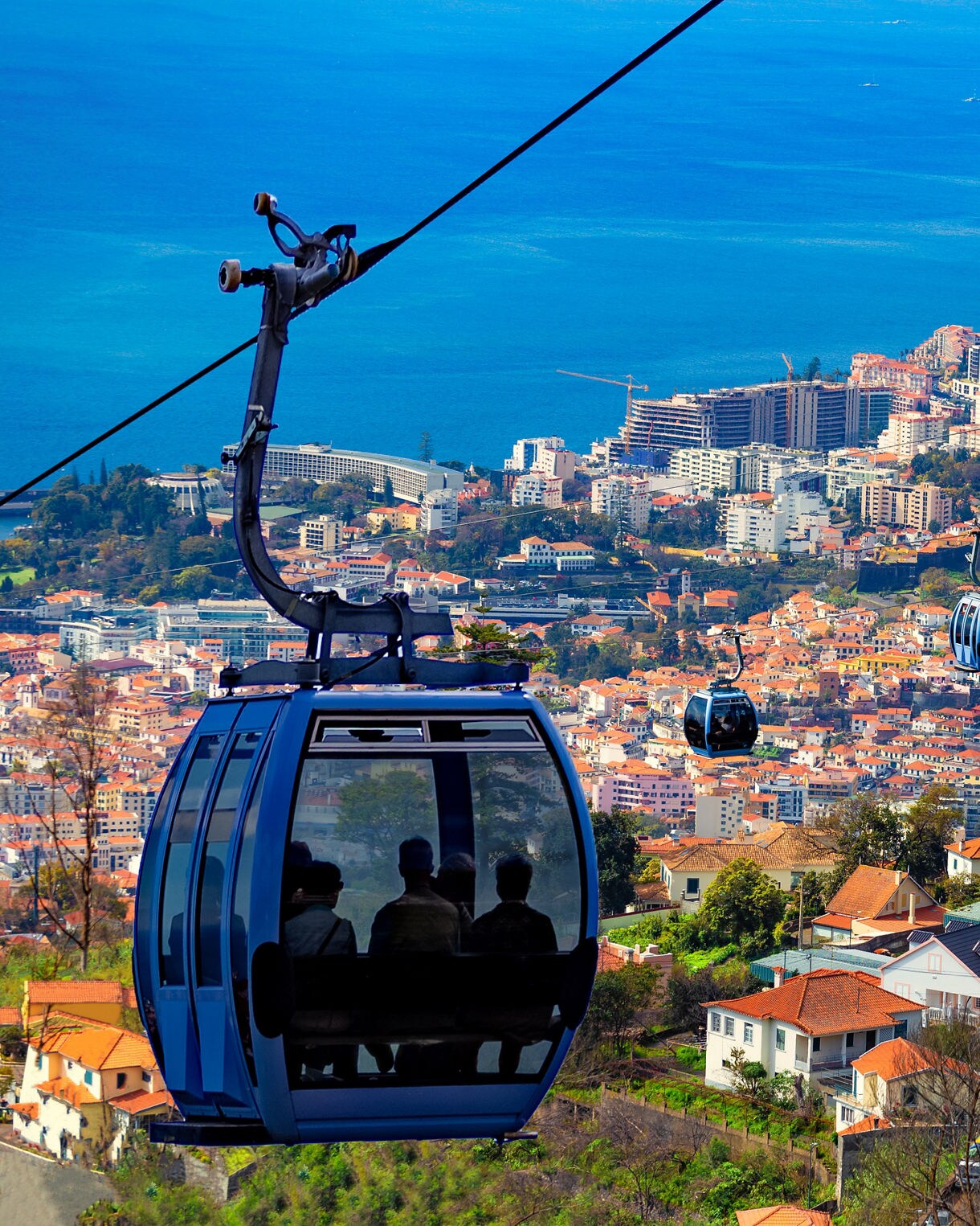 Blue cable cars carrying passengers over the city of Funchal, with red-roofed houses and the Atlantic Ocean in the background.