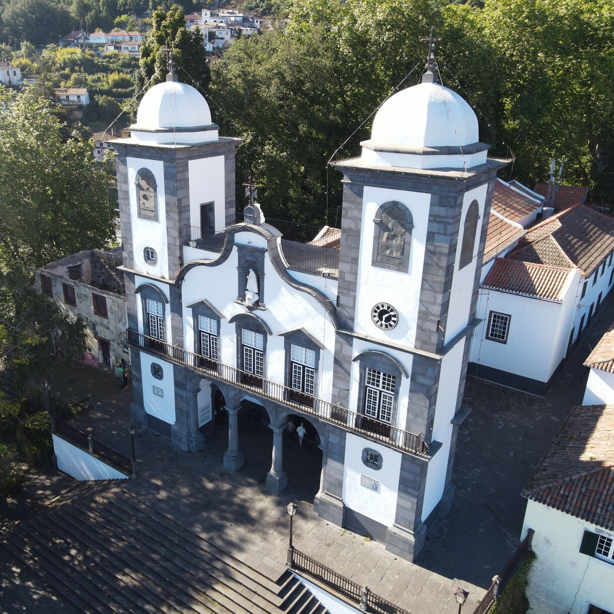 Aerial view of the Church of Our Lady of Monte in Funchal, Madeira, featuring twin white towers with domes, black trim and wide stone steps surrounded by trees.