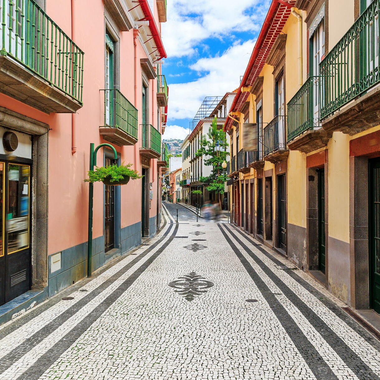 Narrow street in Funchal, Madeira with patterned cobblestone pavement, pastel buildings and wrought-iron balconies.