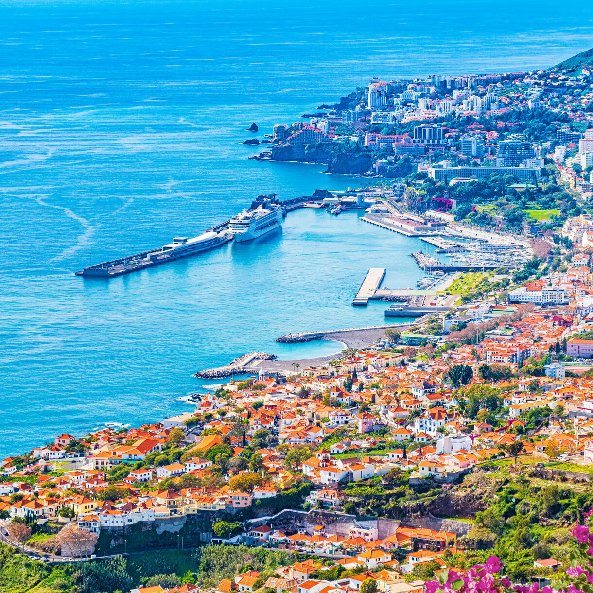 Aerial view of Funchal, Madeira, showing red-roofed houses, cruise ships docked at the harbor and the turquoise Atlantic stretching into the horizon.