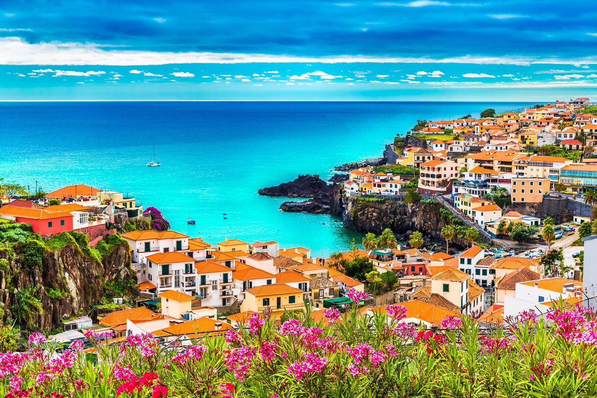 Coastal town in Madeira with white houses and red-tiled roofs perched on cliffs above bright turquoise water, framed by pink flowers in the foreground.