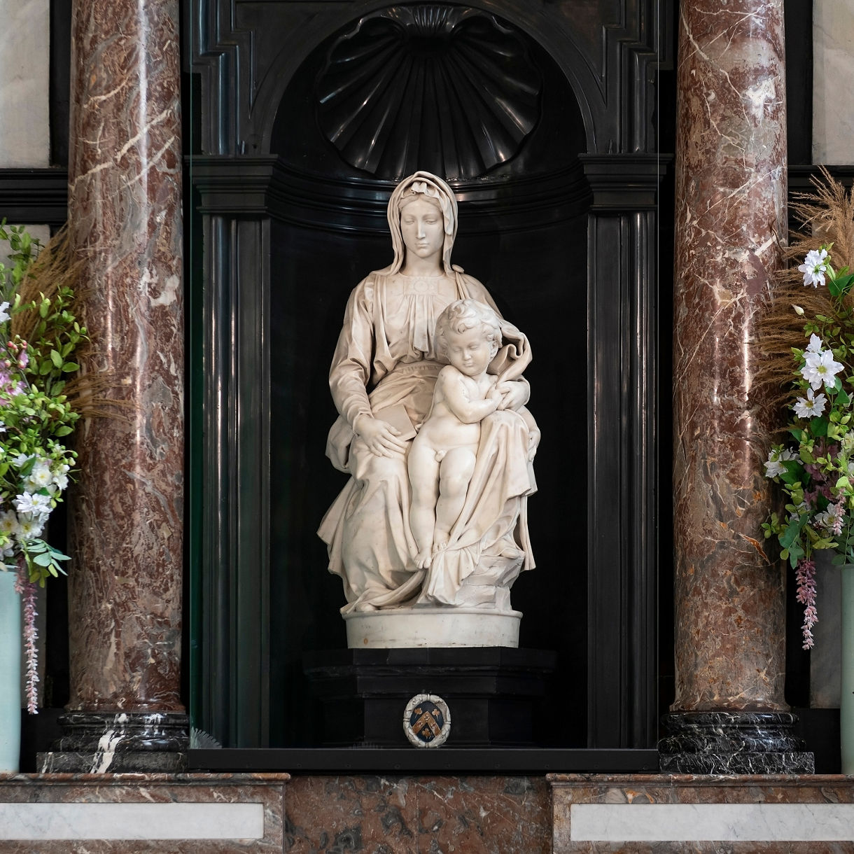 Marble statue of the Madonna and Child by Michelangelo, displayed between dark marble columns with floral arrangements on either side in Bruges, Belgium.