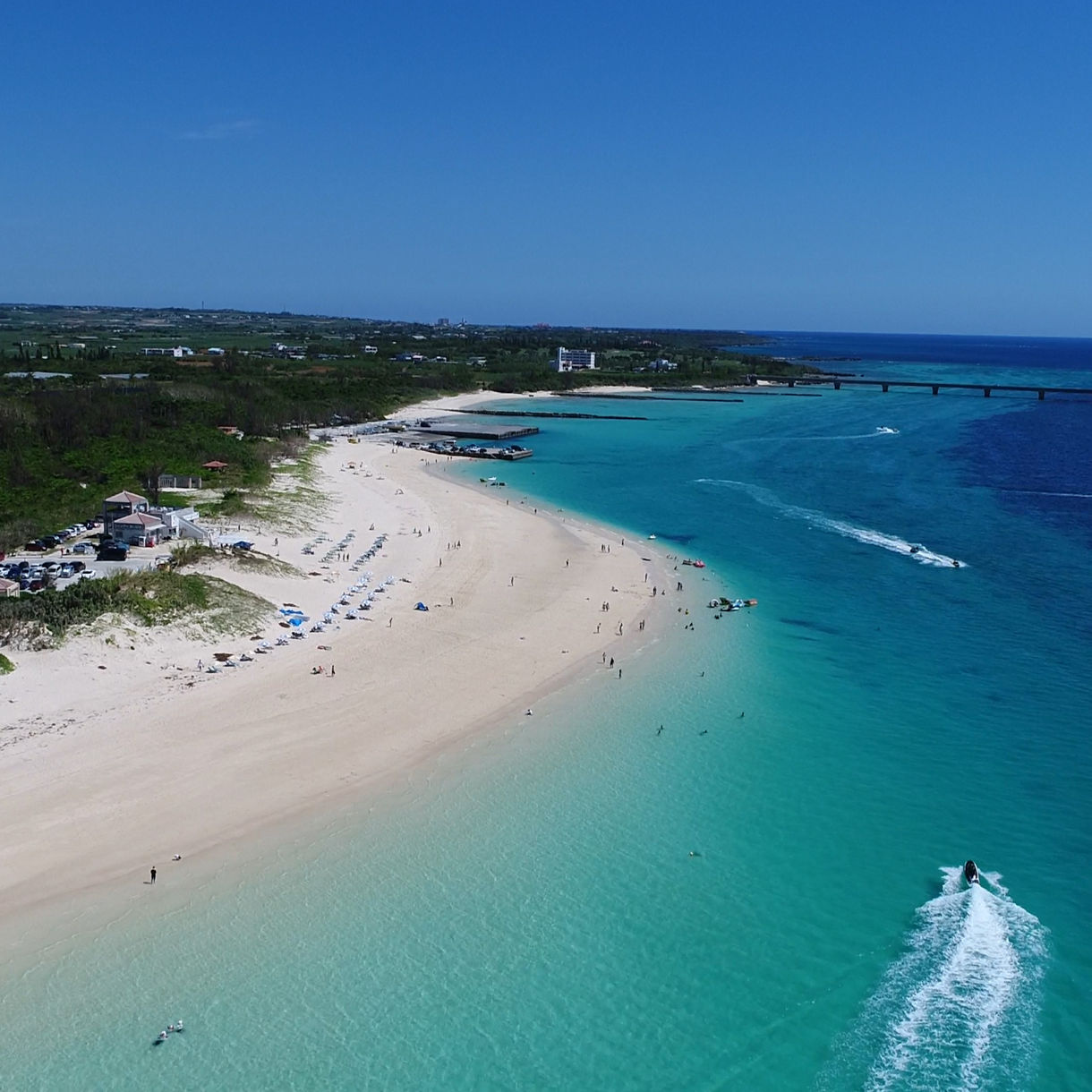 A wide aerial view of Maehama Beach featuring pale sandy shores, clear turquoise water and scattered beachgoers, with a long bridge extending across the ocean in the distance.