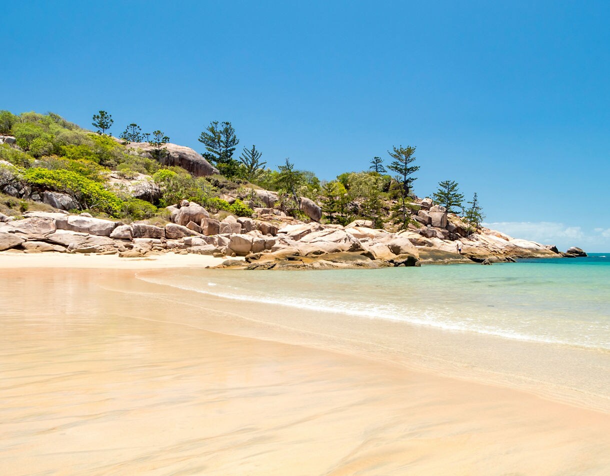 Quiet sandy beach with gentle turquoise waves, rounded granite boulders and pine-topped hills under a bright blue sky.
