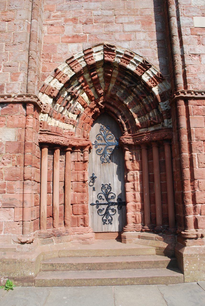 Close-up of St Magnus Cathedral’s arched doorway in Kirkwall, featuring red sandstone carvings, a wooden door and intricate black wrought iron designs.