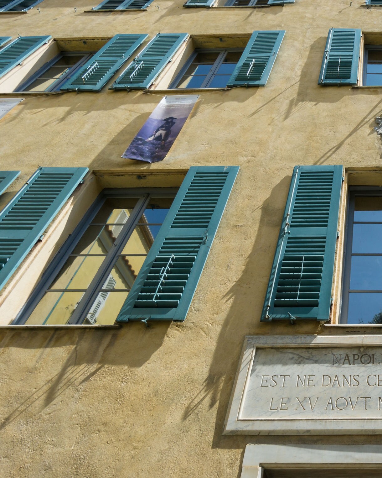 Exterior of Maison Bonaparte in Ajaccio, Corsica, featuring yellow walls, green shutters and a marble plaque marking Napoleon’s birthplace.