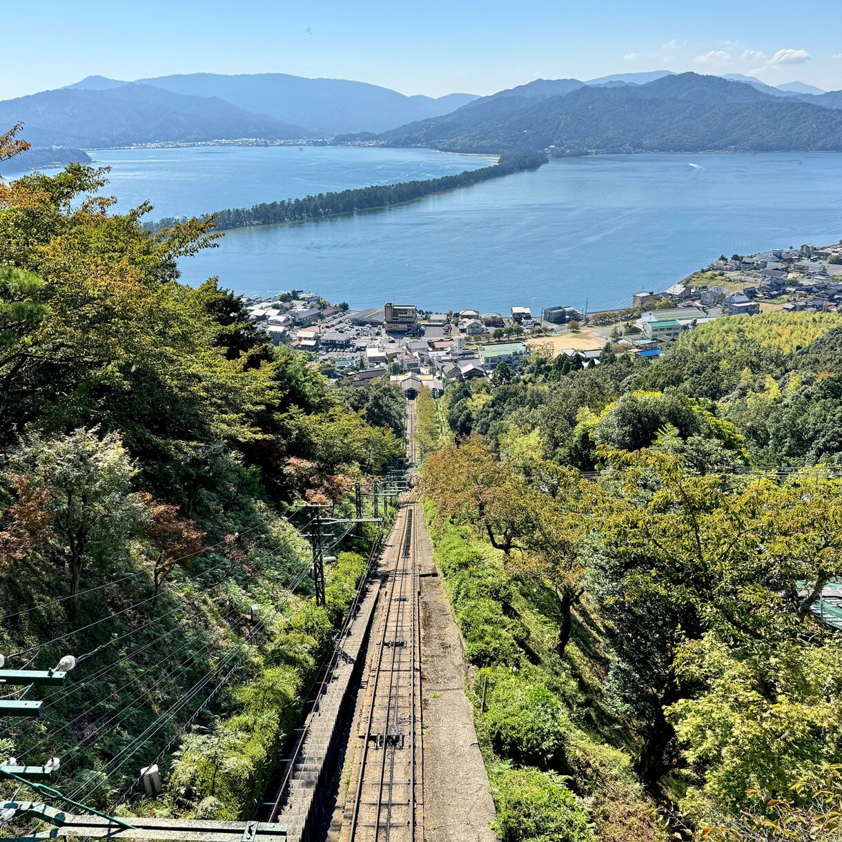 A scenic overlook showing a railway descending a lush green hillside toward a coastal town, with Amanohashidate’s long pine-covered sandbar extending across a calm blue bay and mountains rising in the background.
