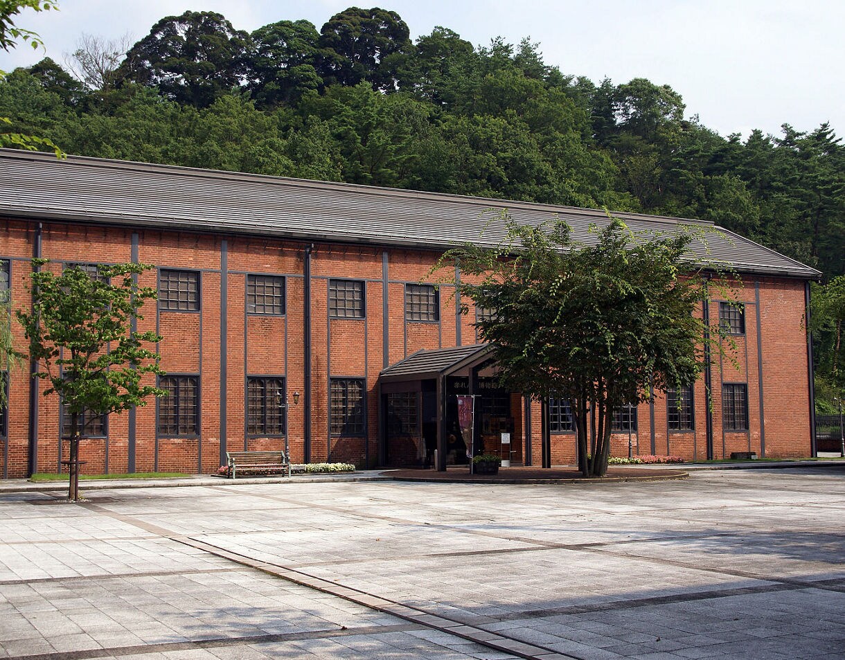 A large red-brick building with tall, grid-paned windows sits beside a tree-lined courtyard at Maizuru’s Brick Museum in Japan.